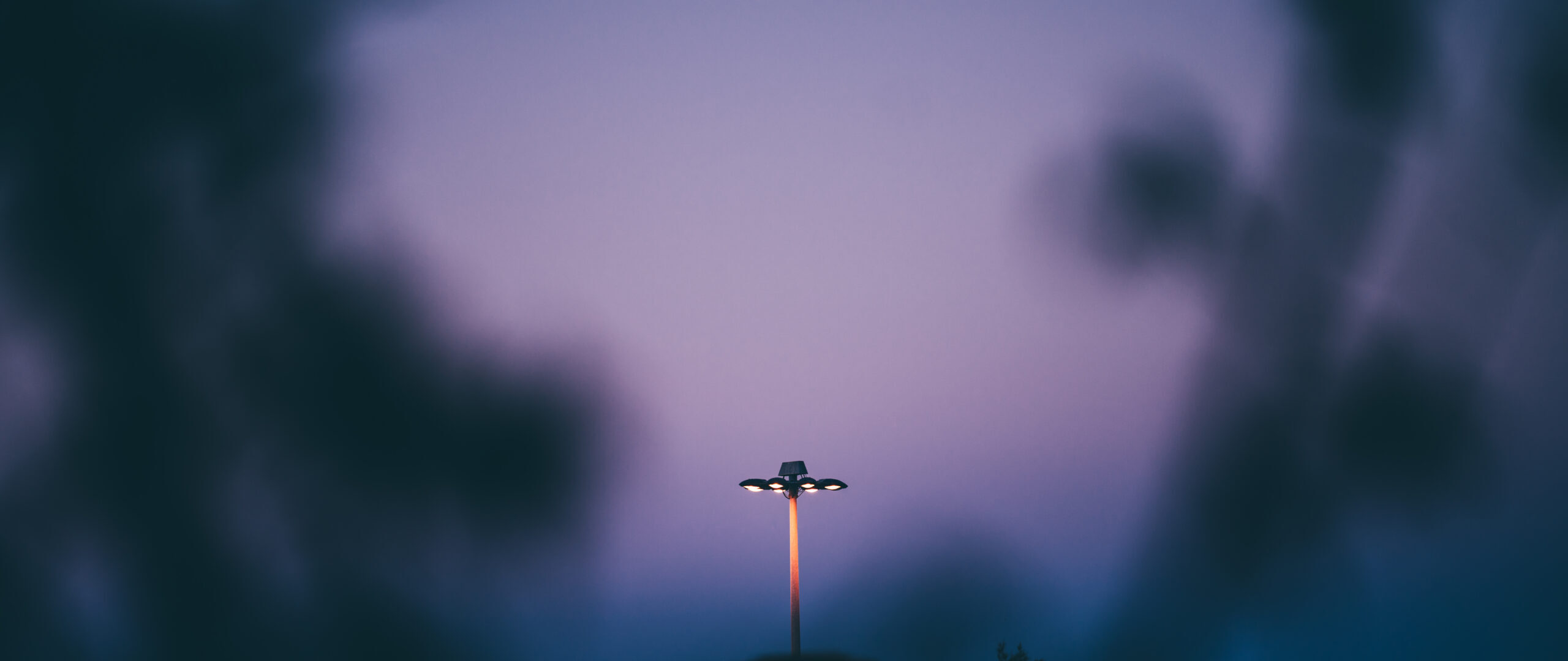 Telephoto shot of a street lamp stands illuminated against a dusk sky, creating a serene and atmospheric scene. Soft shadows frame the image, with a smooth gradient of blue to purple in the background
